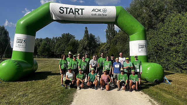 Großer aufblasbarer Startbogen in Grün und Weiß im Freien. Unter dem Bogen steht eine Gruppe von Menschen auf einem hellen Weg, alle tragen leuchtend grüne T‑Shirts mit weißen Aufdrucken; viele haben schwarze oder dunkle kurze Hosen an, einige tragen Kappen. Der Bogen ist links und rechts wie zwei dicke grüne Säulen, oben mit einem grünen Querbalken verbunden. Auf dem rechten, weißen Mittelteil steht senkrecht groß „START“, darunter „AOK“ und darunter kleiner „aok.de/lauf“. Auf den weißen Feldern an den unteren Außenseiten des Bogens steht jeweils „FIRMEN LAUF“ mit AOK‑Logo.