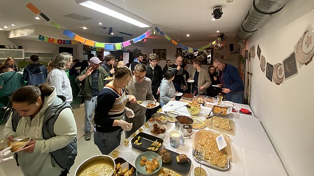 Im Cafe Carisma stehen viele Menschen in einem bunt dekoriertem Raum. Auf einem langen Tisch stehen viele unterschiedliche Gerichte