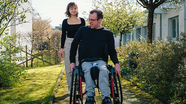 Ein Mann, der einen Rollstuhl nutzt und eine junge Frau sind draußen im Freien auf einem Weg.