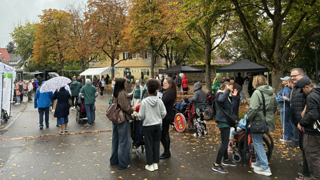 Herbstliche Parkszene mit vielen Menschen auf einem Platz, teilweise mit Rollstühlen und Hilfsmitteln.   Der Boden ist nass, überall liegen gelbe und braune Blätter, es wirkt wie ein kühler, regnerischer Tag.   Im Hintergrund stehen Reihen hoher Bäume mit herbstlich gefärbtem Laub, dahinter ein hellgelbes Gebäude mit mehreren Fenstern.   Links sammeln sich Menschen, einige mit Kinderwagen, eine Person hält einen bunten Regenschirm.  