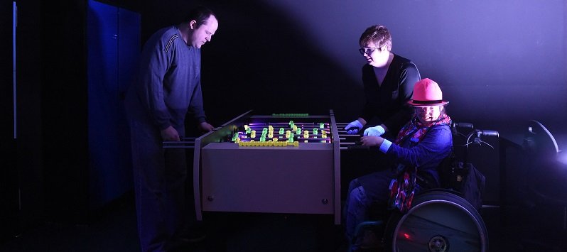 Drei Personen spielen im Dunkeln an einem leuchtenden Tischkicker
