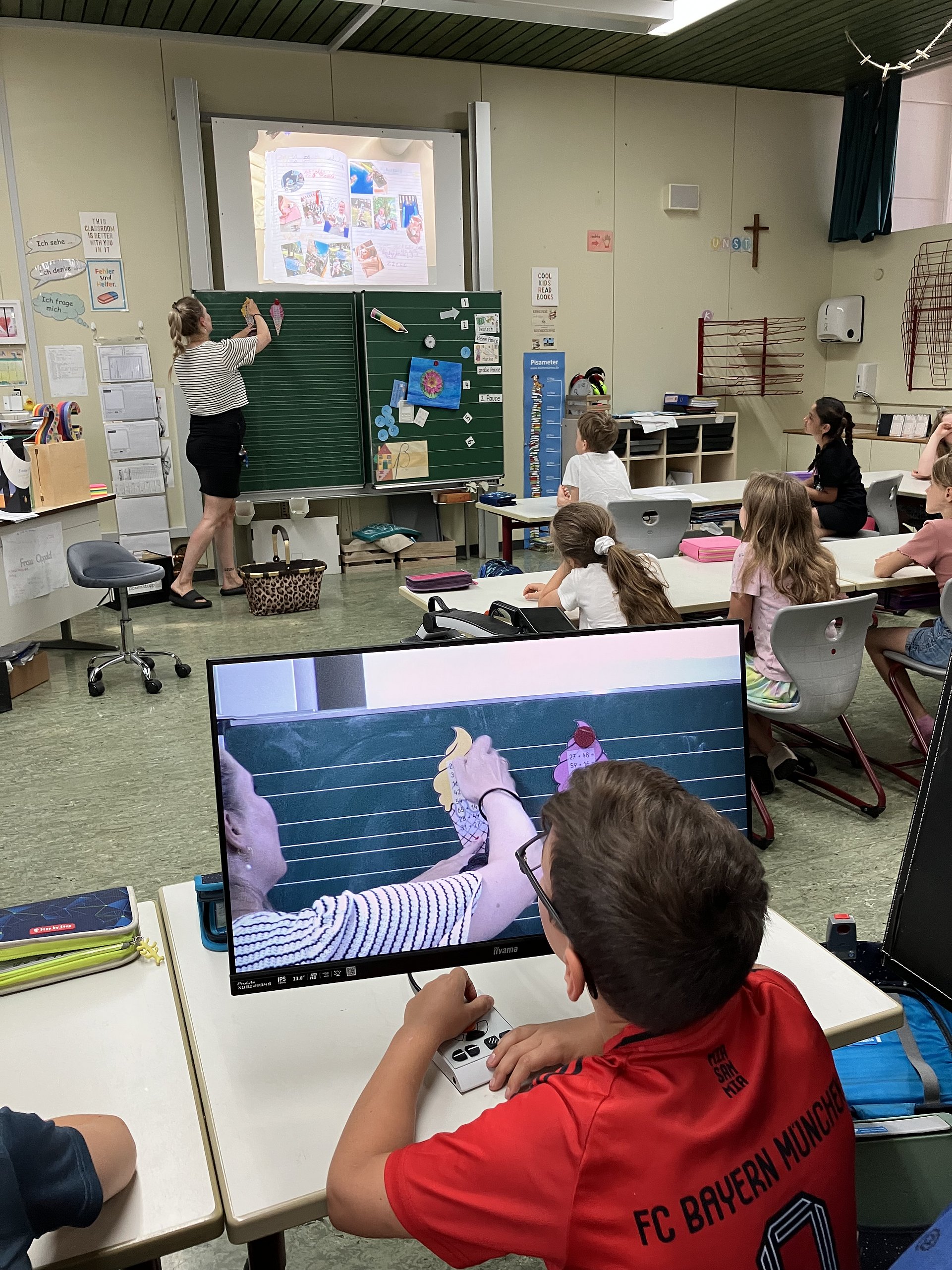 Schülerinnen und Schüler in einem Klassenzimmer. Die Lehrerin steht vorne an der Tafel und schreibt etwas darauf. Ein Schüler in einem roten T-Shirt und einer Brille sitzt vor einem Bildschirm und sieht in der Vergrößerung, was die Lehrerin an die Tafel schreibt