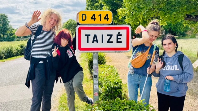 Eine Frau steht mit weiteren drei jungen Frauen auf der Straße neben einem Straßenschild. Auf diesem ist der Name einer französischen Stadt zu lesen: Taisé