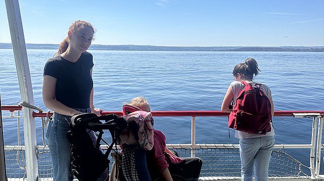 Eine junge Frau steht an deck eines Schiffes. Es fährt über den Bodensee und die Sonne scheint. Ihre Hände liegen auf dem Lenker eines Rollstuhles, in dem eine Klientin sitzt und auf den See schaut. Rechts neben ihnen steht eine weitere Frau. Sie lehnt mit ihrer Körpervorderseite an der Rehling. Sie trägt einen Rucksack. 