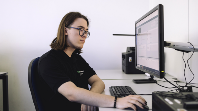 An einem Schreibtisch sitzt ein junger Mann. Er trägt ein schwarzes T-Shirt, Schulter lange braune glatte Haare und eine Schwarz umrandete Brille. Mit seiner rechten Hand bedient er eine Computermaus. Er sitzt vor einem angeschalteten Computer und arbeitet daran.