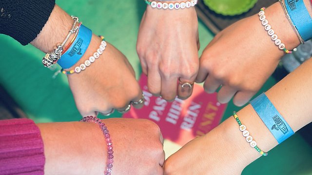 Fünf, zu Fäusten geballte Hände treffen sich in der Mitte des Bildes. an jedem Handgelenk hängen Perlenarmbänder