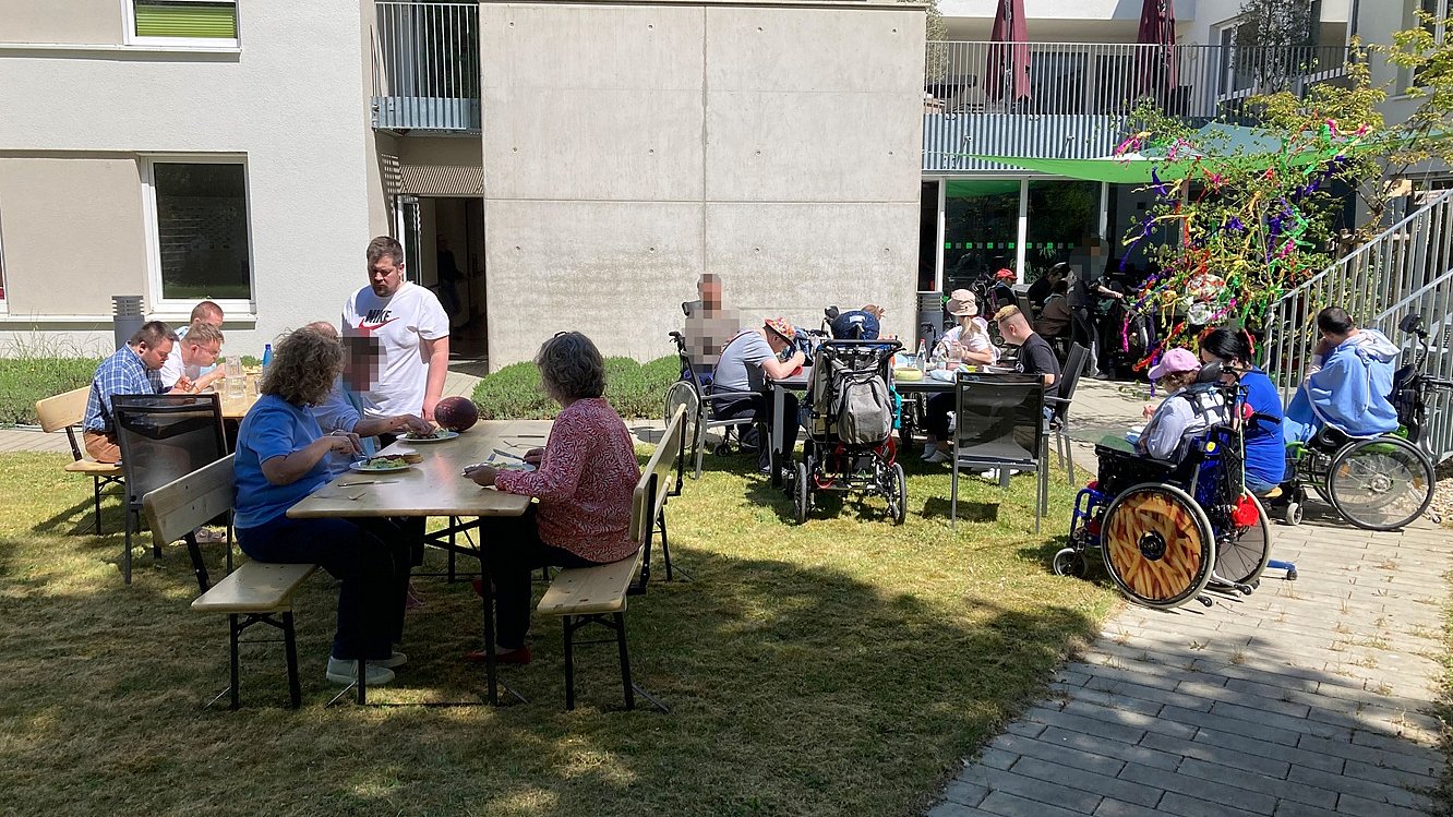 Auf einer Wiese vor einem Haus stehen Tische und Stühle, auf denen Menschen sitzen, sich unterhalten und essen. Links sitzen zwei Personen im Rollstuhl vor einem Treppenaufgang.