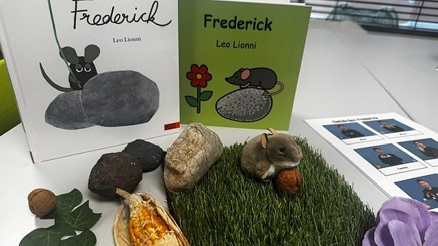 Auf einem Tisch stehen zwei Bilderbücher die eine Maus zeigen und den Titel Frederick tragen. Vor den Büchern liegen Steine, Blätter, ein Maiskolben und ein rechteckiges Stück Kunstrasen. Darauf sitzt eine kleine Plüschmaus, die eine Walnuss hält. 