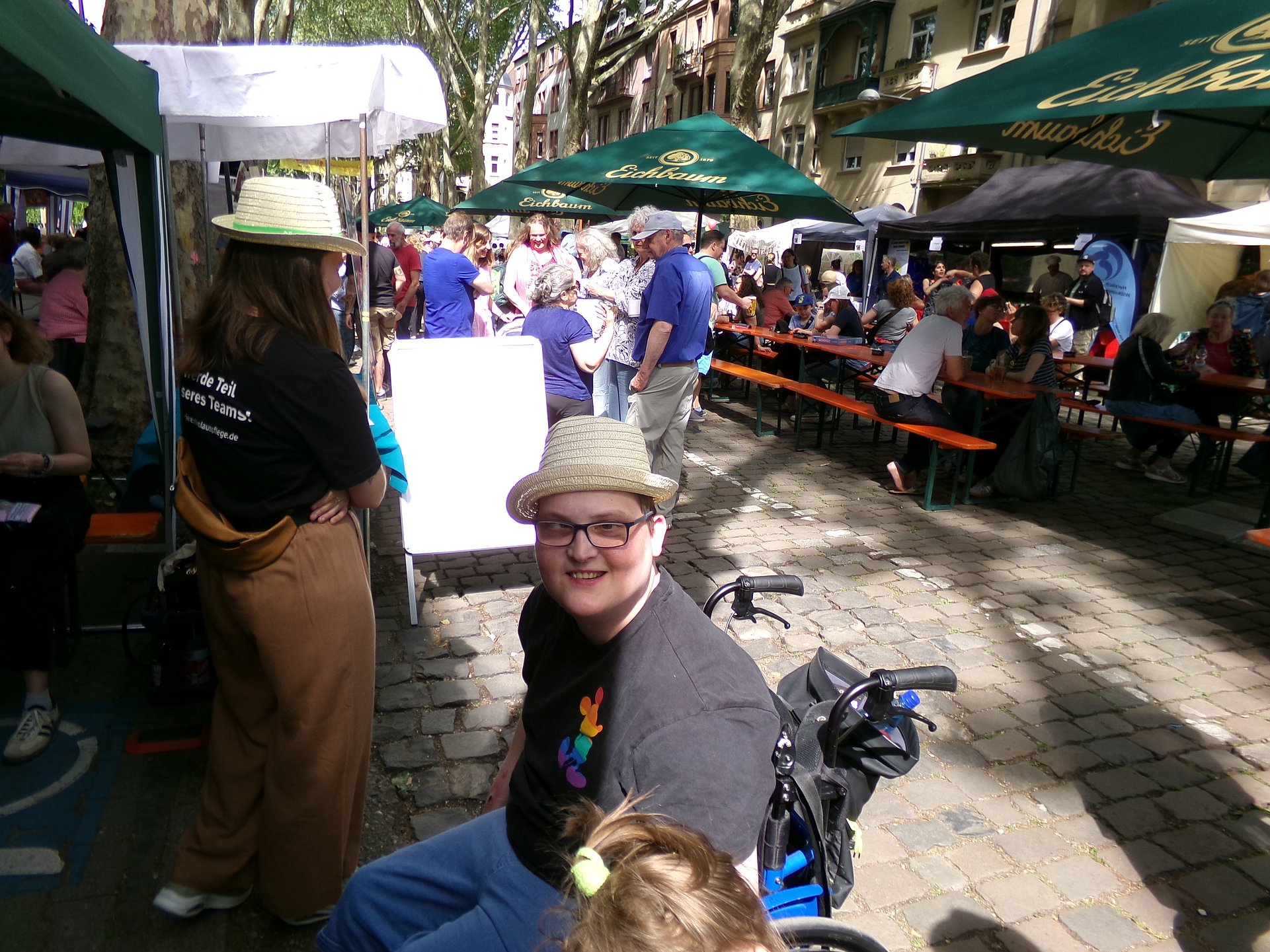 Auf dem Strassenfest sind viele Stände und Menschen. Die Sonne scheint. Mitten im Bild ist ein Mann im Rollstuhl zu sehen. Er trägt eine Brille und ein Sonnennhut und lacht fröhlich