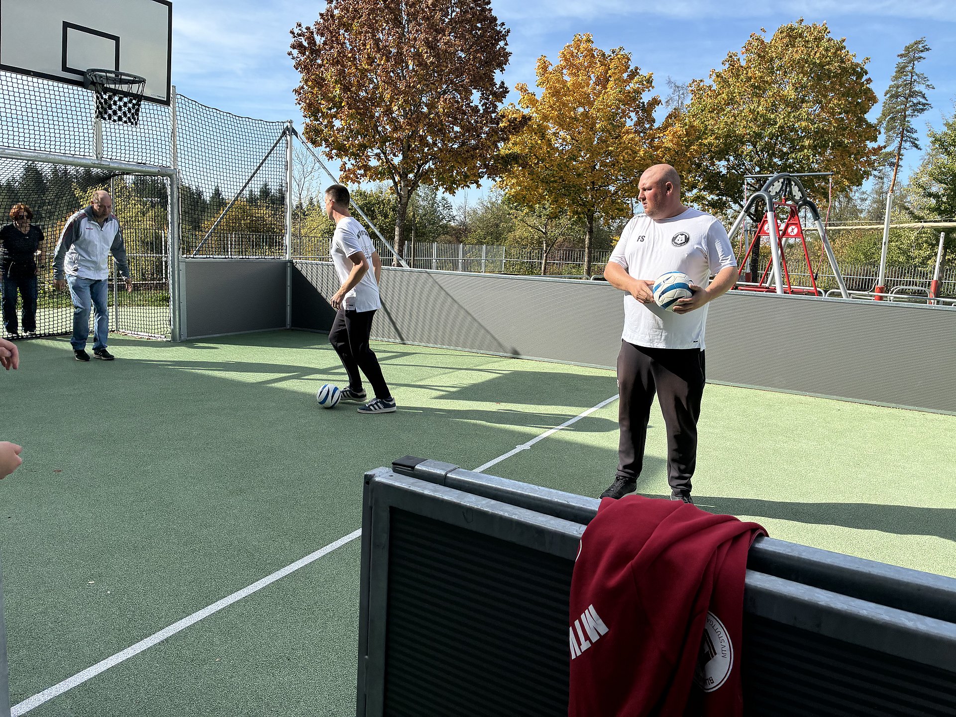 Drei Personen auf einem kleinen Sportplatz mit Kunstrasen und Basketballkorb.