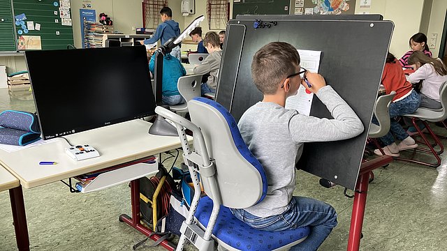 In einem Klassenzimmer sitzen Schülerinnen und Schüler. Im Vordergrund sitzt ein Schüler auf einem Schreibtischstuhl an einer Tafel. Darauf ist ein Blatt Papier befestigt. Er sitzt mit dem Gesicht ganz nah daran und schreibt mit seiner rechten Hand etwas darauf. Neben ihm auf dem Schreibtisch steht ein großer schwarzer Bildschirm