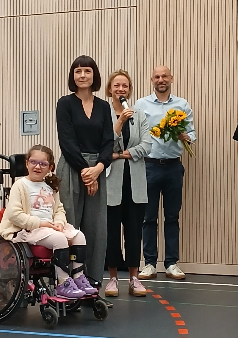 Zwei Frauen und ein Mann stehen nebeneinander. Die mittlere Frau hält ein Mikrofon. Der Mann rechts von ihr hält einen Blumenstrauß. ganz links im Bild sitzt eine Schülerin im Rollstuhl und lacht freundlich