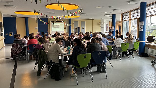 In einem hellen großen Raum sitzen viele Personen gemeinsam an zusammengestellten Tischen und unterhalten sich. An der Decke sind bunte Wimpel aufgehängt