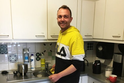 Ein junger Mann mit gelb-schwarzem Sweatshirt steht in einer Küche und lächelt