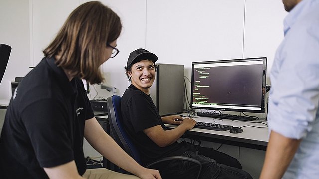 Ein Jugendlicher sitzt an einem Schreibtisch auf dem ein Computer steht. Er dreht sich zur Kamera und lacht. Links neben ihm sitzt ein weiterer Jugendlicher und ist ihm zugewand.
