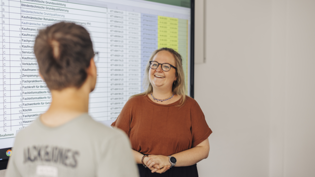 Im Vordergrund sieht man einen jungen Mann von hinten. Vor ihm steht eine Frau die freundlich lächelt. Links neben ihnen an der Wand wird eine Excelltabelle an die Wand vergrößert projeziert