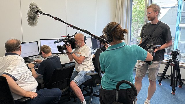 Vier Personen sitzen und stehen in einem Unterrichtsraum. Links sitzt ein Mann in Jeans und weißem T-Shirt und blickt vor sich auf einen Bildschirm auf dem Tisch. Rechts daneben sitzt ein junger Mann, er trägt eine schwarze Brille und tippt auf einer Tastatur, vor ihm stehen Bildschirme auf dem Tisch. Rechts daneben sitzt ein Mann und zielt mit einer großen Kamera auf die beiden Männer neben ihm. Rechts dahinter steht eine weitere Person und hält ein Mikrofon an einem langen Stab über die Personen vor ihr.