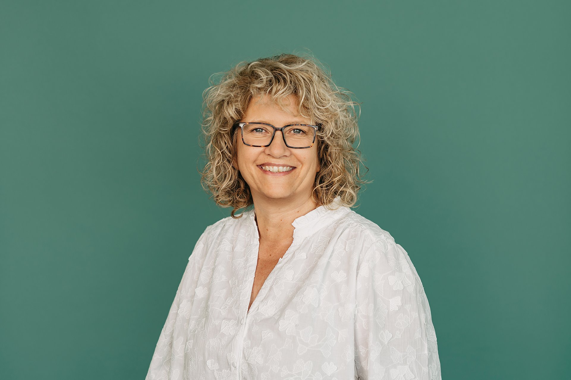 Eine Frau vor einem grünem hintergrund. Sie trägt eine weiße Bluse und eine dunkel umrandete Brille. Sie hat blonde lockige schulterlange Haare und lächelt freundlich