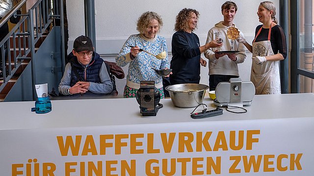 Mitarbeitende und Teilnehmervertretende stehen hinter einem langen Tisch. Dieser ist mit einer weißen Tischdecke gedeckt. Darauf stehen Schüsseln und ein Waffeleisen. Eine Mitarbeiterin schöpft Teig mit einer Schöpfkelle aus einer Schüssel. 