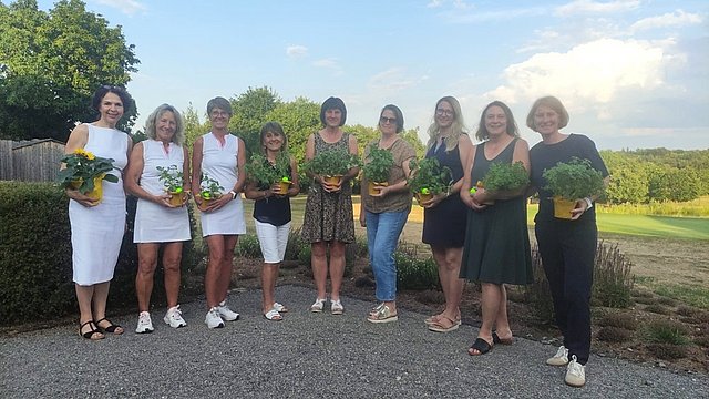 Ein Foto mit neun Frauen nebeneinanderstehend vor einer Wiese mit Bäumen. Sie tragen weiße und dunkle, schicke Kleidung und halten kleine Blumengestecke in der Hand. Sie lächeln alle in die Kamera. 