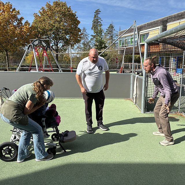 Drei Erwachsene und ein Kind auf einem kleinen Fußballfeld mit Tor. Links steht eine Frau mit hellbraunen, schulterlangen Haaren, die nach hinten zusammengebunden sind. Sie trägt ein olivgrünes T‑Shirt, eine beige Schürze oder Weste und eine hellblaue Jeans mit weißen Sportschuhen. Vor ihr ist ein spezieller Kinderrollstuhl mit drei Rädern und seitlicher Stützkonstruktion. In diesem sitzt ein Kind mit pinkfarbenen Polstern am Sitz und an den Fußstützen. Die Frau beugt den Oberkörper nach vorn über das Kind und hält einen weiß‑blauen Fußball direkt vor das Gesicht oder den Oberkörper des Kindes, als ob sie beim Schießen oder Passen helfen würde.