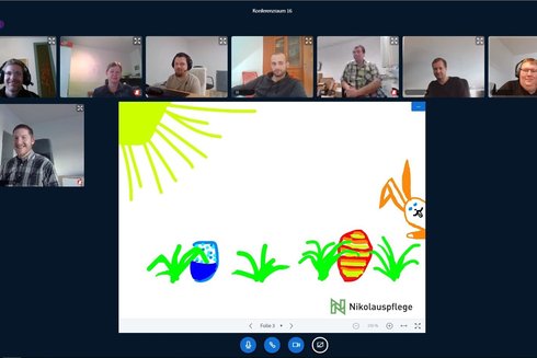 Ein Bildschirm zeigt eine Videokonferenz mit acht Personen. In der Mitte ein gemaltes Osterbild.