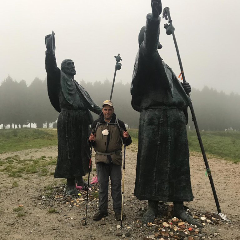 Andreas Natterer steht mit Wanderstöcken vor zwei Pilgerstatuen 