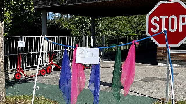 Ein kleiner Zielbogen aus weißen Stangen über einem grünen Weg. Zwischen den Stangen sind mehrere bunte, durchscheinende Tücher aufgehängt: von links nach rechts ein blaues, ein pinkes, ein blaues, ein grünes und ein pinkes Tuch. Oberhalb der Tücher hängt in der Mitte ein weißes Blatt Papier mit handgeschriebener Aufschrift, darauf steht: „ZIEL“.