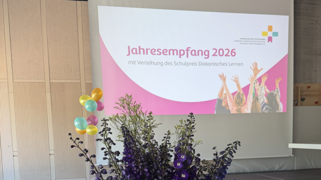 Großer, bunter Blumenstrauß in einer Turnhalle. Der Strauß steht auf einem schwarzen Podest und besteht aus vielen unterschiedlichen Blumen: kräftig orangefarbene, gelbe, rote und pinke Blüten im unteren Bereich, dazwischen kleinere violette und blaue Blüten, oben heraus ragen lange, schlanke, dunkelviolette Blütenrispen. Unten herum sind reichlich grüne Blätter und Zweige als Umrandung angeordnet. Im Hintergrund hängt eine große Leinwand. Darauf ist eine Präsentationsfolie zu sehen. Darauf steht in großer Schrift: „Jahresempfang 2026“. Darunter in kleinerer Schrift: „mit Verleihung des Schulsiegels Diakonisches Lernen“.  Links an der Leinwand hängen mehrere bunte Luftballons in Gelb, Orange, Blau und Pink.