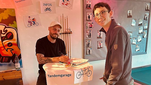 Ein junger Mann steht an einem Informationsstand. Dahinter steht noch ein Mann. Beide lächeln. An dem Stand wird über Tandemfahrten informiert