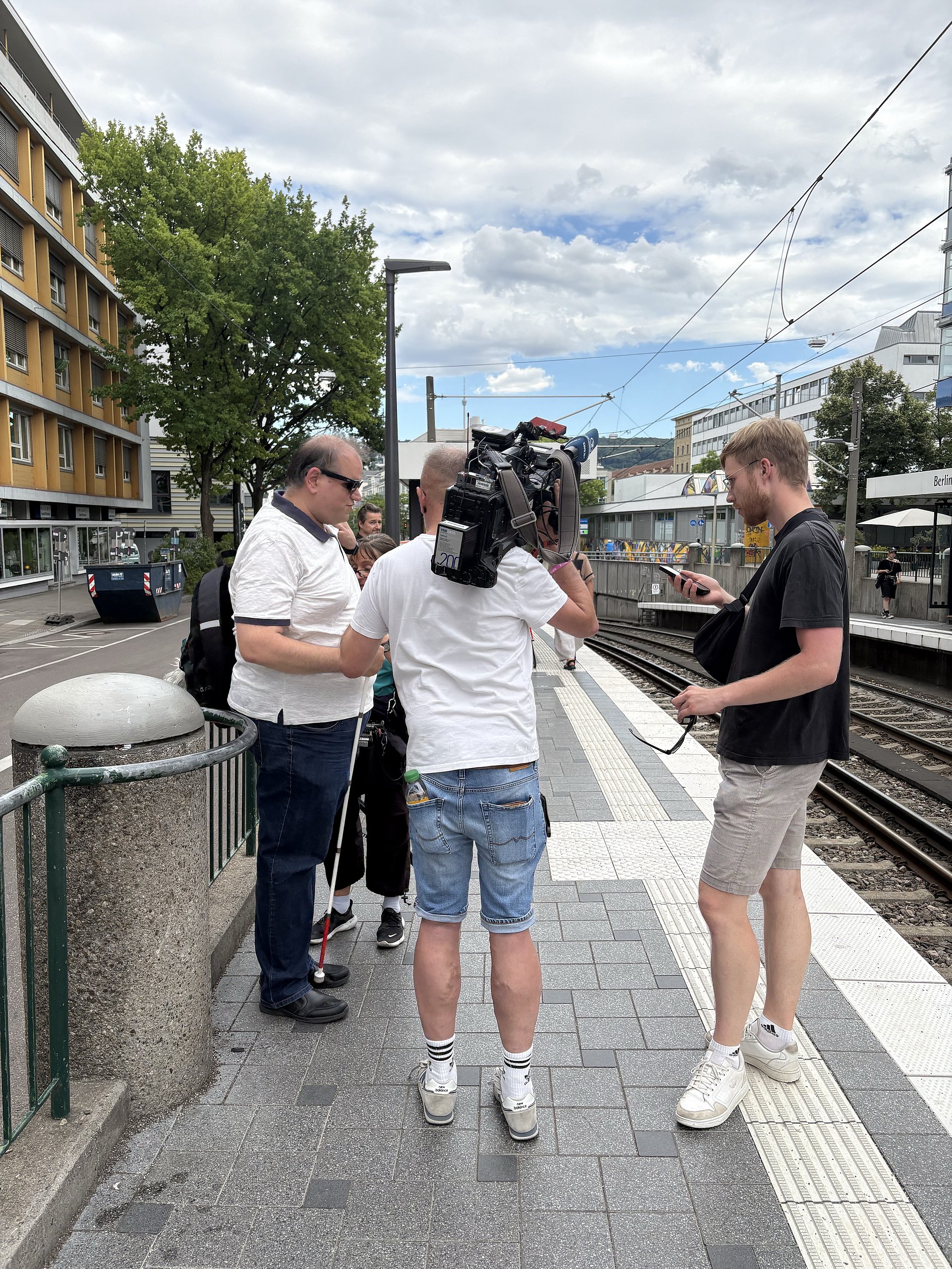 Vier Personen auf einem U-Bahngleis in Stuttgart. Links ein Mann in hellen Shorts und schwarzem T-Shirt, der sich von der Kamera wegdreht. Dahinter eine Person mit einer großen Kamera auf der Schulter. Rechts daneben ein Mann in Jeans, weißem T-Shirt und weißem Langstock in der Hand, er trägt eine schwarze Brille. Rechts vor ihm eine Person in schwarzer Hose und türkisfarbenem T-Shirt, die ein Mikrofon an einem langen Stab in der Hand hält. Im Hintergrund sind Straßen und Häuser zu erkennen, an einer Häuserwand ist ein großes buntes Graffiti.
