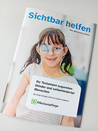 Auf einem hellgrauem Tisch liegt ein Heft. Darauf ist ein lächelndes Mädchen abgebildet mit einer Brill. Das linke Brillenglas ist mit einem bunten Pflaster abgeklebt