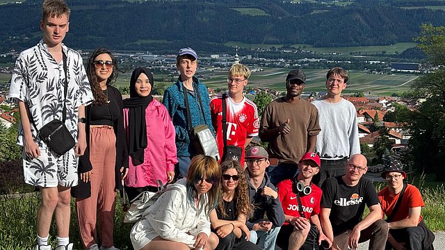 Eine größere Gruppe junger Menschen sitzt und steht im Freien vor einer Berglandschaft.   Im Vordergrund sitzt eine Reihe von Personen auf einem niedrigen Mäuerchen oder Steinrand. Alle wirken entspannt und posieren nah beieinander für ein Gruppenfoto. Im Hintergrund erstreckt sich ein weitläufiges Tal mit Feldern, einem Ort mit orangefarbenen Dächern und hohen grünen Bergen dahinter.