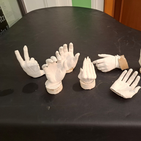 Auf einem dunklen Tisch liegen acht weiße Hände aus Gips in verschiedenen Formen