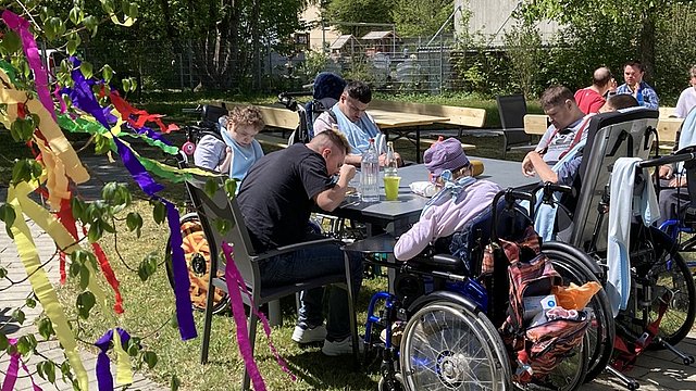 Auf einer Wiese stehen Tische und Stühle, an denen Menschen sitzen und essen. Einige davon sitzen im Rollstuhl. Im Hintergrund sind Bäume und Büsche zu sehen, links wehen bunte Papierfahnen ins Bild hinein. 