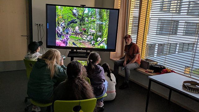 Schülerinnen und Schüler sitzen im Klassenzimmer vor einem großen Bildschirm und sehen sich einen Film an. 