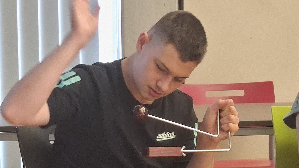 Ein junger Mann sitzt auf einem Stuhl und bedient eine kleine Triangel. Er trägt ein schwarzes T Shirt mit einem weißen „adidas“-Logo auf der Brust und hat kurz geschnittene, dunkle Haare. Sein Kopf ist leicht nach vorn und unten geneigt, der Blick geht konzentriert auf das Instrument in seiner rechten Hand. Das Instrument besteht unten aus einem flachen rechteckigen Holzblock, aus dem ein gebogener Metallbügel nach oben führt. Oben am Metallbügel ist eine dunkle Kugel befestigt, die wie eine kleine Holzkugel wirkt. Die linke Hand des Mannes ist angehoben, als würde er gleich auf die Kugel oder den Bügel schlagen oder eine Bewegung ausführen. Im Hintergrund stehen einfache Stühle, einer mit roter Rückenlehne.