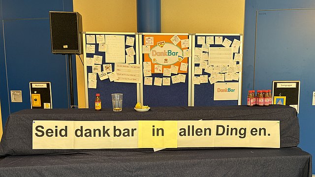 Auf einem Kirchaltar stehen Pinwände, an denen viele beschriftete Zettel angebracht sind. Unten auf dem Altar hängt ein großes Banner auf dem steht: Seid dankbar in allen Dingen. Links auf dem Altar steht ein Lautsprecher
