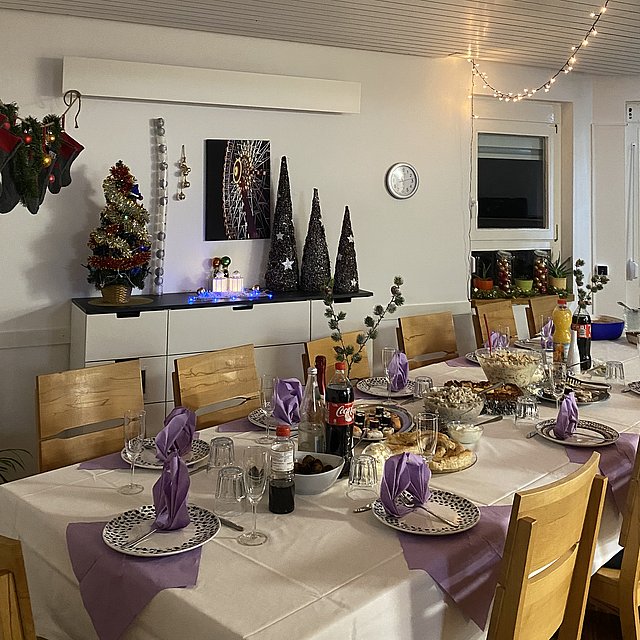 Ein gedeckter Tisch mit einer weißen Tischdecke steht in einem Zimmer. Darauf stehen Gläser, Teller, Besteck, Servietten, Blumen und Kerzen. 