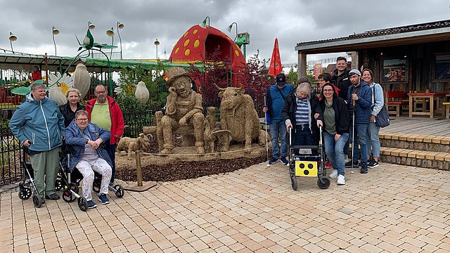 Gruppenfoto vor einer großen Fantasiefigur aus Stroh. In der Mitte stehen und sitzen mehrere Erwachsene in zwei Reihen, einige mit Rollstuhl oder Rollator. Vorne sitzen und stehen vier Personen dicht beieinander, alle in Freizeitkleidung mit Jacken; die Farben sind überwiegend Blau, Schwarz und Dunkelgrau. Einer hält einen weißen Langstock, ein anderer stützt sich auf einen Rollator, an dem vorn ein gelber Kasten mit schwarzen Punkten befestigt ist. Hinter ihnen stehen weitere Personen, teils Arm in Arm; Jacken in Hellblau, Rot, Beige und anderen Farben. Die Menschen haben unterschiedliche Haarfarben von grau über blond bis dunkel und wirken überwiegend älter bzw. mittelalten Alters. Direkt hinter der Gruppe stehen mehrere große Figuren aus goldbraunem Stroh oder Heu, die wie sitzende und liegende Fantasietiere oder Bären gestaltet sind. Eine der Figuren hat ein Gesicht mit Brille und Hut. Daneben liegen kleinere Tierfiguren aus demselben Material. Die Figuren stehen in einem Beet mit dunklem Rindenmulch, das mit einem Seil abgegrenzt ist.