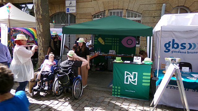 Auf einem Straßenfest im Freien Steht ein Informationsstand der Nikolauspflege. Ein Pavillon und darunter ein Stehtisch mit einer Stoffverkleidung. Alles in Dunkelgrün. Vor dem Stand stehen zwei Mitarbeiterinnen und zwei Klienten die im Rollstuhl sitzen. Sie Sonne scheint.