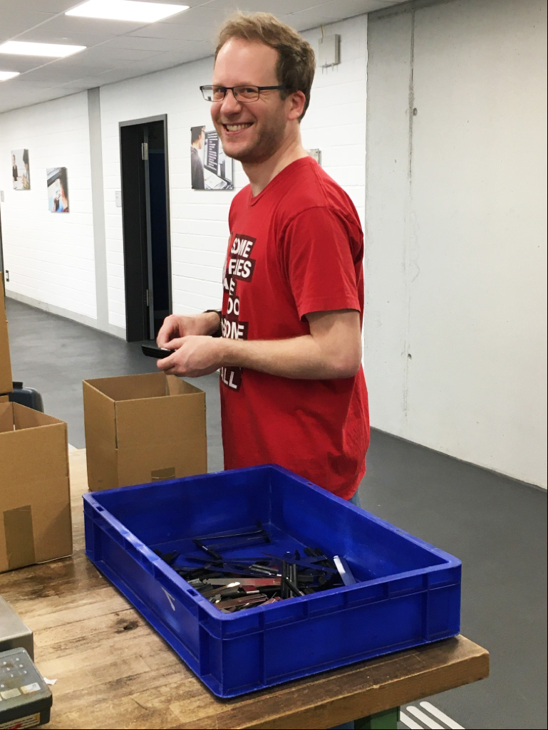 Ein junger Mann mit rotem T-Shirt steht in einer Werkstatt und lacht.