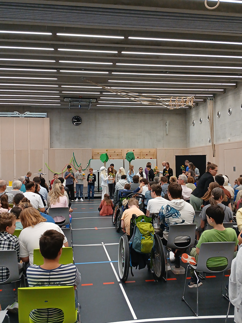 In der Turnhalle sitzen viele Menschen auf grünen Stühlen. Vorne ist eine Bühne zu sehen