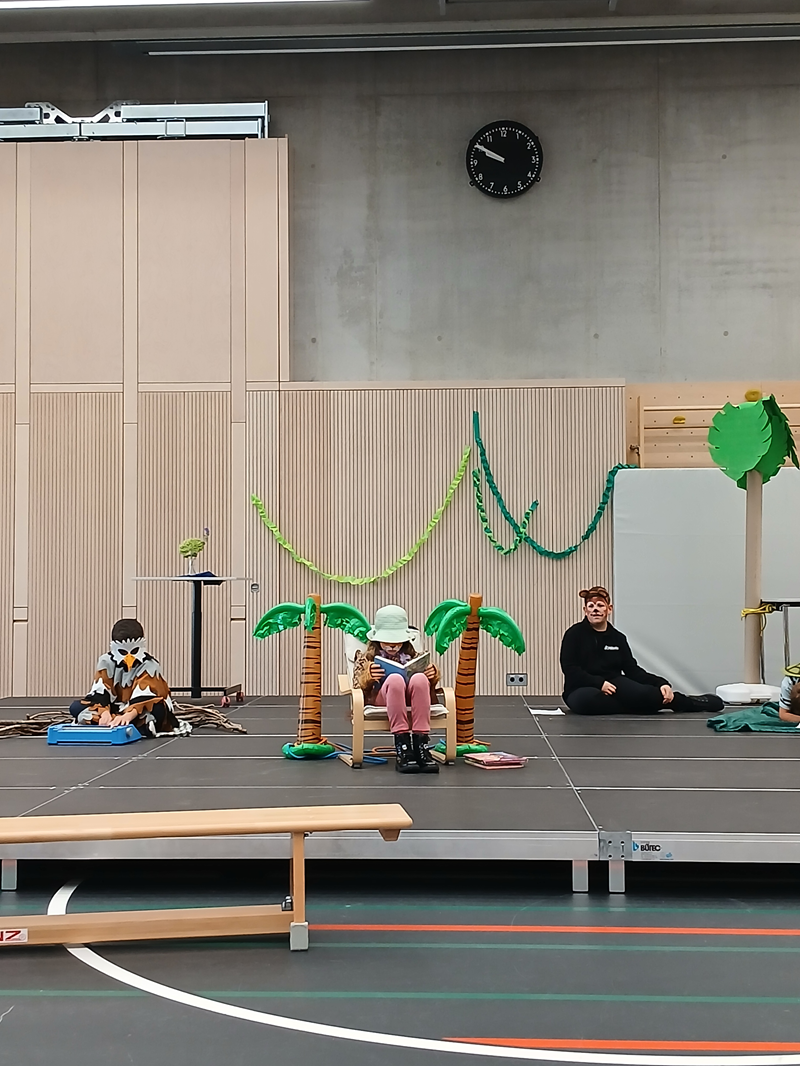Auf einer Bühne in der Sporthalle sind drei verkleidete Kinder. Zwei Dekopalmen und grüne Girlanden schmücken die Bühne