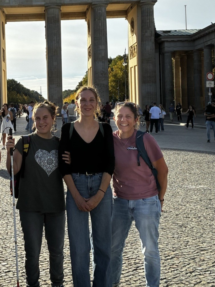 Drei Personen, die vor dem Brandenburger Tor stehen. In der Mitte steht eine große Person mit nach hinten gebundenen Haaren, schwarzem langärmligem Oberteil mit V-Ausschnitt und blauer Jeans, die Hände sind vor dem Körper gefaltet und die Person lächelt. Links daneben steht eine Person mit dunkelbraunen zusammengebundenen Haaren, einem dunkelgrauen T Shirt mit einem großen, hellen Herzmotiv auf der Brust und einer dunklen Hose; in der rechten Hand hält die Person einen weißen Langstock, der nach unten zeigt. Rechts steht eine Person mit lockigen zu einem Dutt gebundenen Haaren, einem rosafarbenen T Shirt, blauer Jeans und einem dunklen Rucksack über der rechten Schulter, ebenfalls lächelnd. Die drei stehen dicht beieinander auf einem gepflasterten Platz und haben die Arme zum Teil umeinander gelegt. Im Hintergrund ist das Brandenburger Tor mit seinen hohen Säulen und dem Durchgang zu sehen. Hinter dem Trio gehen und stehen viele weitere Menschen auf dem Platz, einige machen Fotos.