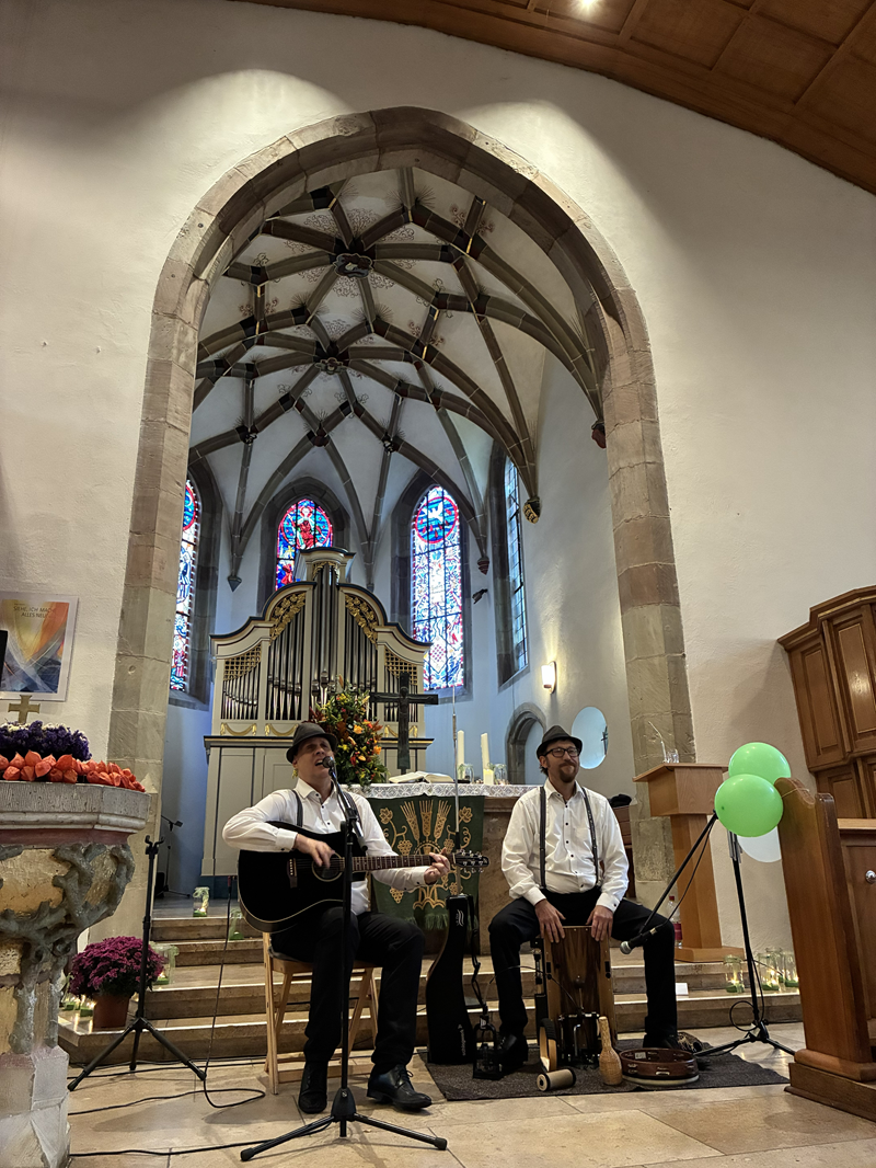 Ein Kircheninnenraum mit zwei Musikern im Altarbereich.Im Hintergrund steht ein großer gotischer Chorraum mit hohem Spitzbogen und dunklem hölzernem Gewölbe. Dahinter sind mehrere hohe, schmale Kirchenfenster mit farbigen Bleiglasmotiven zu erkennen. Vor den Fenstern steht eine große Pfeifenorgel mit hellen Holz- und Metallpfeifen, daneben ein üppiger Blumenstrauß in kräftigen Farben.