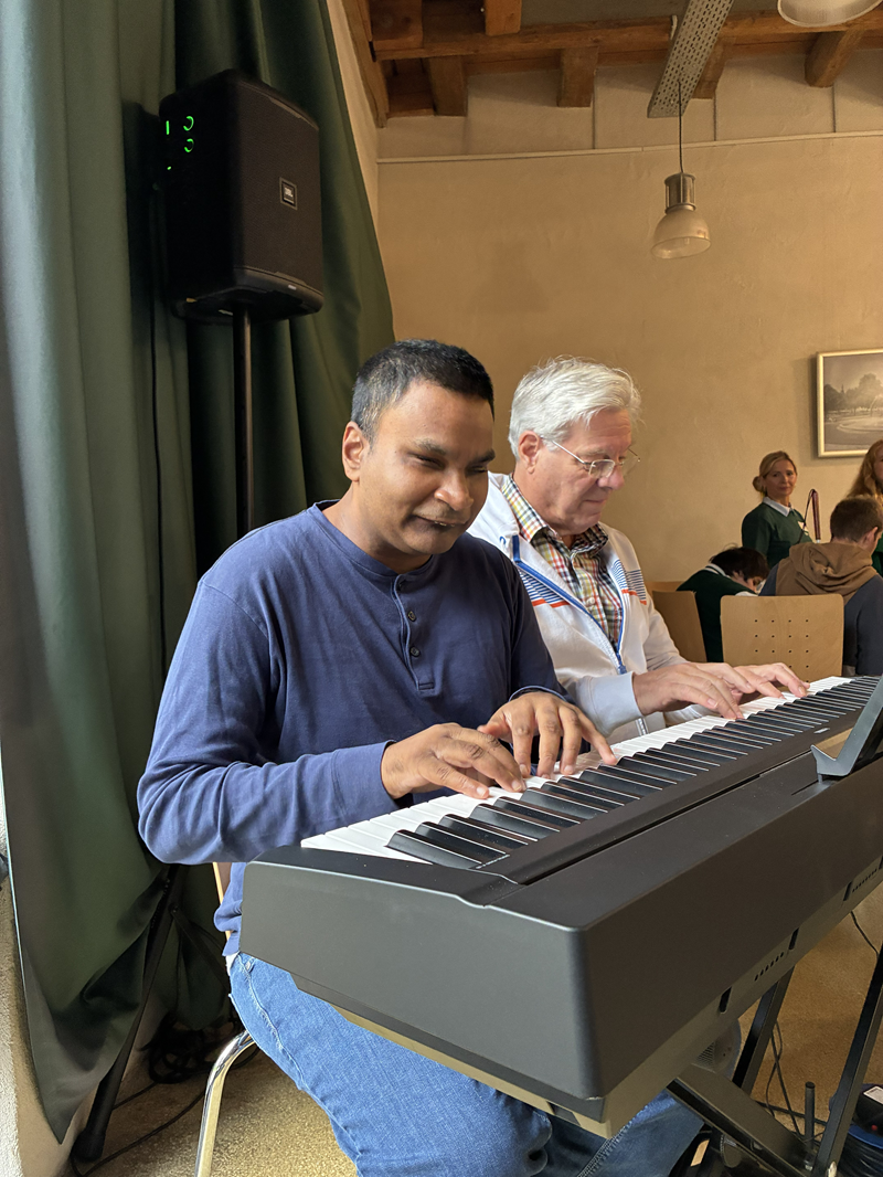 Zwei Männer spielen gemeinsam an einem elektronischen Keyboard. Vorne sitzt ein Mann mit mittelbrauner Haut, kurzen dunkelgrauen Haaren und einem dunkelblauen, langärmeligen Henley-Shirt sowie einer hellblauen Jeans. Seine Hände liegen auf den Tasten des Keyboards, der Mund ist leicht geöffnet, der Gesichtsausdruck wirkt konzentriert und zugleich gelöst. Direkt hinter ihm sitzt ein älterer Mann mit heller Haut, weißen Haaren und Brille. Er trägt ein hellblaues Hemd mit einem bunt karierten Kragen und ein helles Namensband um den Hals. Auch seine Hände liegen auf der Klaviatur, als würden beide vierhändig spielen.