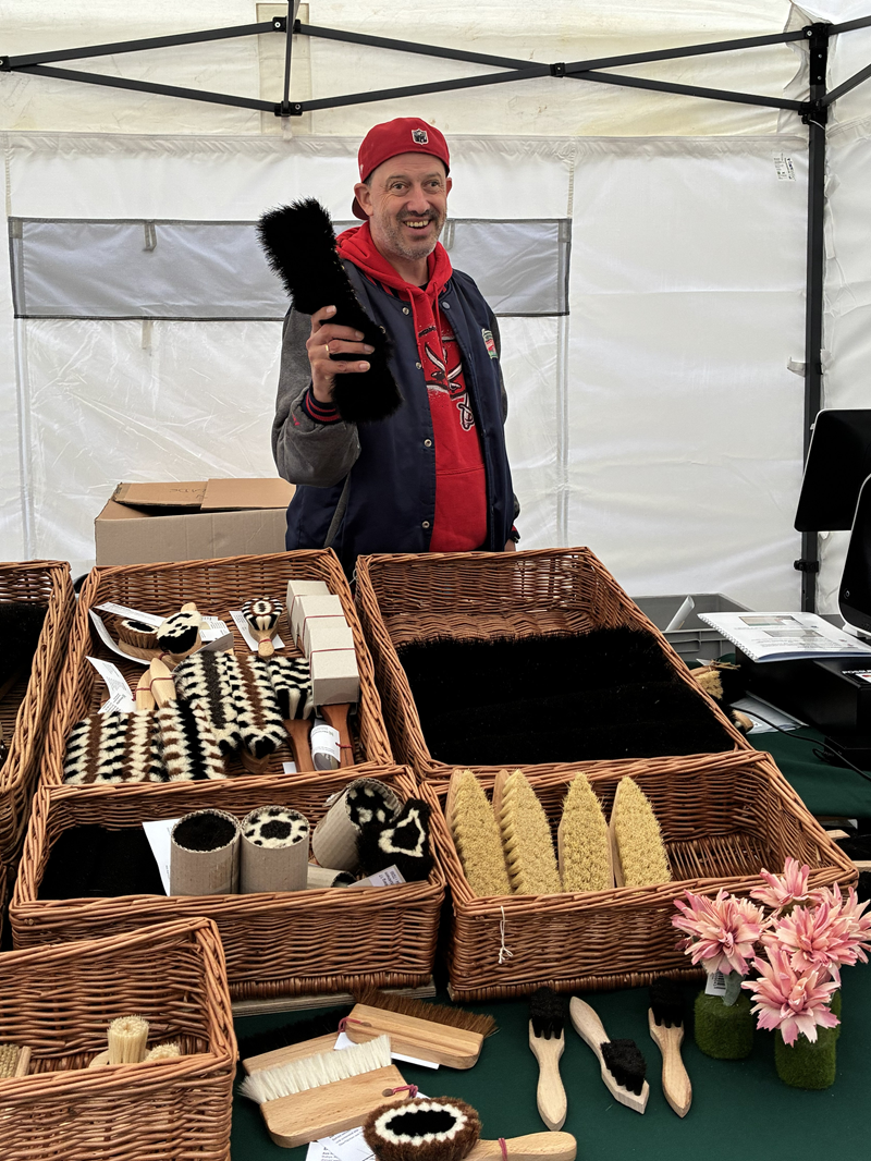 Ein Marktstand mit Korbwaren und Bürsten. In der oberen Bildhälfte steht unter einem gelb‑weißen Pavillon ein Mann mittleren Alters. Er trägt eine rote Baseballkappe rückwärts, einen roten Kapuzenpullover mit Aufdruck, darüber eine dunkle Collegejacke mit grauen Ärmeln. Er lächelt breit und hält in der rechten Hand eine schwarze, flaschenförmige Bürste nach vorne. Hinter ihm hängt die helle Seitenwand des Zelts mit einem waagerechten Fensterstreifen, daneben steht ein schwarzer Stuhl und Kartons liegen auf einem Tisch.