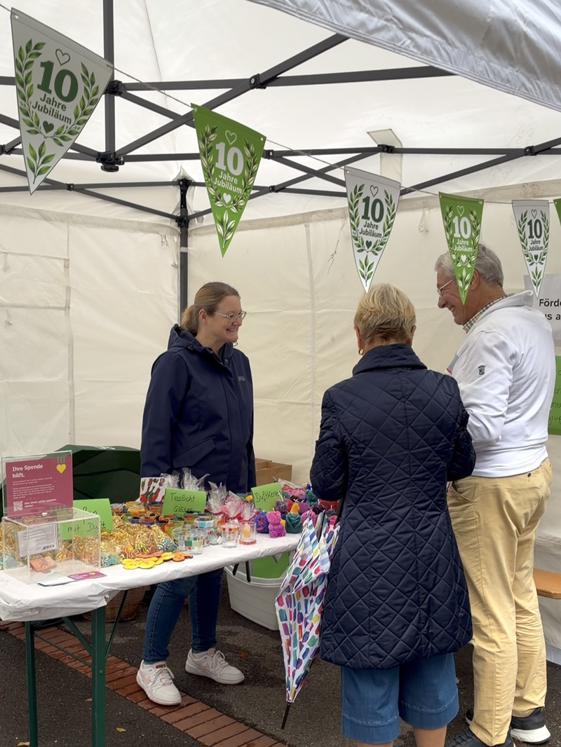 Marktstand unter einem Pavillon mit Jubiläumsdekoration. Unter einem weißen Zelt stehen drei Erwachsene zusammen an einem Klapptisch mit vielen kleinen bunten Artikeln, vermutlich Selbstgebasteltes oder Süßigkeiten in Tütchen und Schalen. Links steht eine Frau mit heller Haut, dunkelblauer Regenjacke, blauen Jeans und weißen Turnschuhen, sie lächelt in Richtung der beiden anderen Personen. In der Mitte steht eine Frau mit heller Haut, dunkelblauer, gesteppter Jacke, knielangen blauen Hosen und beigen Schuhen, sie trägt eine bunte Einkaufstasche an der rechten Seite und ist dem Tisch zugewandt. Rechts steht ein Mann mit heller Haut, weißem Pullover und hellbeigen Hosen, er beugt sich etwas zum Tisch und wirkt, als würde er sich unterhalten oder etwas erklären.