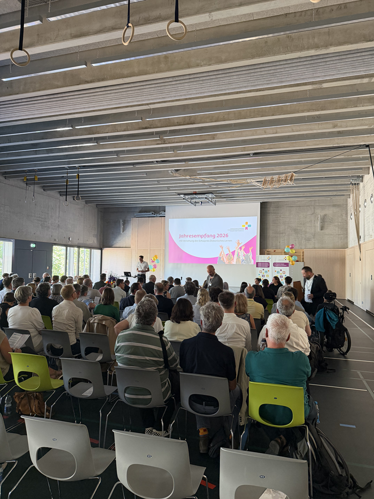Im Vordergrund des Fotos sitzen viele Menschen auf Plastikstühlen in grau und grün. Die Personen blicken nach vorn zu einer Bühne. Vorne im Raum ist eine Leinwand mit einer farbigen Präsentation zu sehen. Auf der Präsentation steht „Jahresempfang 2026“. Neben der Leinwand sind zwei Roll-Ups sowie bunte Luftballons. Der Boden der Turnhalle ist grau, von der Decke hängen einige Turnringe herab. Vorne auf der Bühne steht ein Mann an einem Rednerpult.